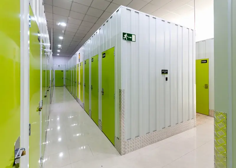 Pasillo de trasteros con puertas verdes y señal de salida de emergencia en Keepy Ciudad Jardín, Málaga